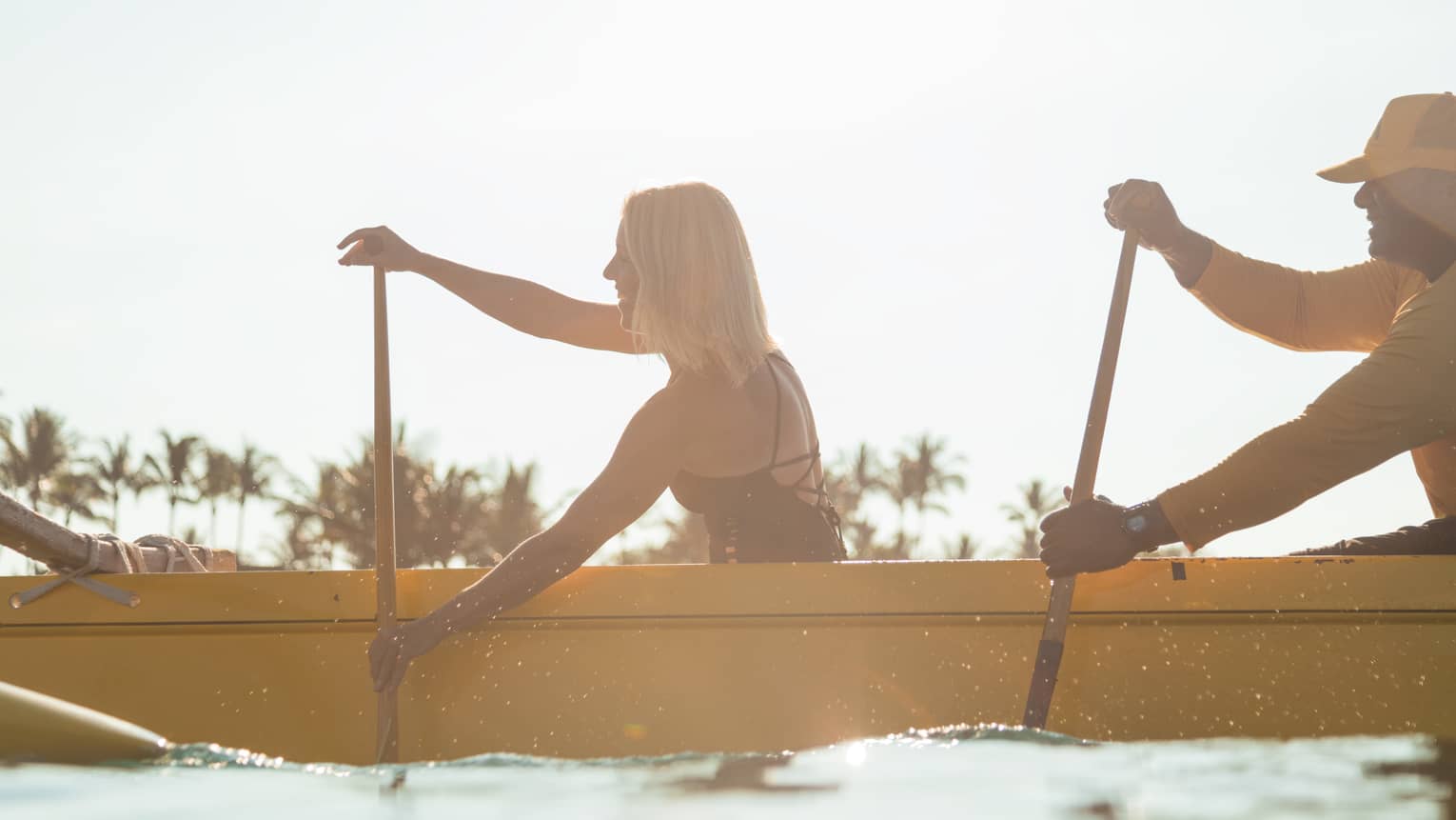 Side view silhouette of woman in swimsuit, man in yellow uniform paddling in yellow canoe, palm trees in background