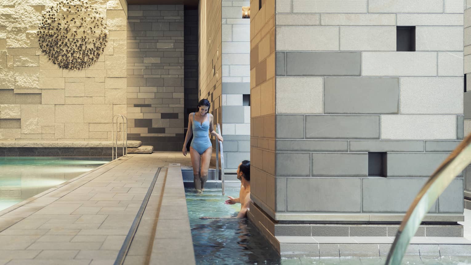 A guest enters a pool via the stairs to meet another guest who is half-hidden by a large grey-tiled column in the foreground.