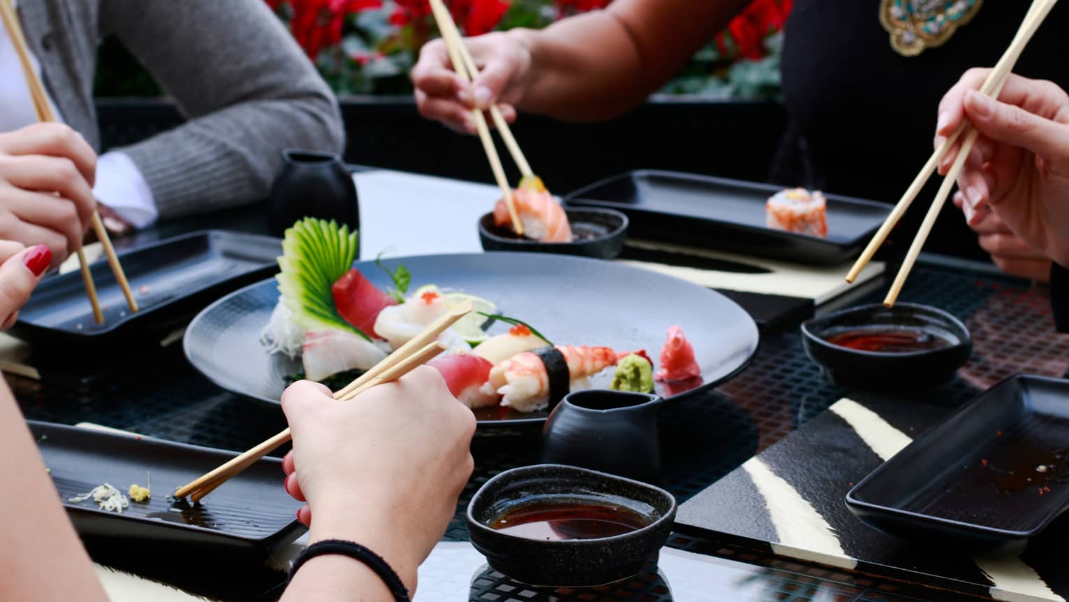 Close-up of four sets of hands holding chopsticks around black plate with sushi rolls, bowls of soy sauce