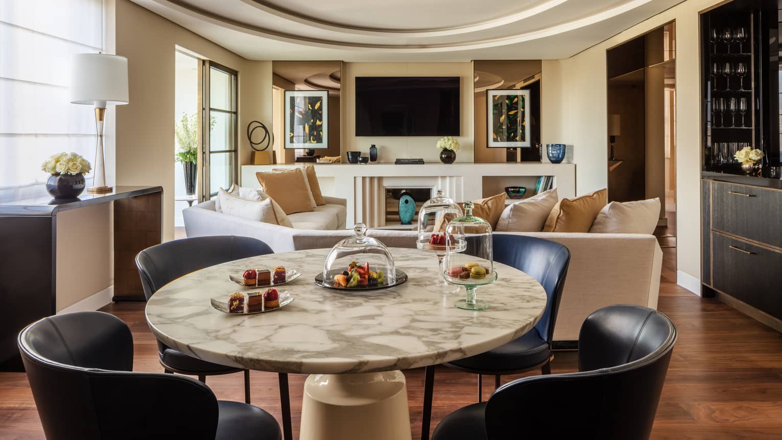 A suite with a round stone table with black chairs, a beige sofa, a television and large windows.