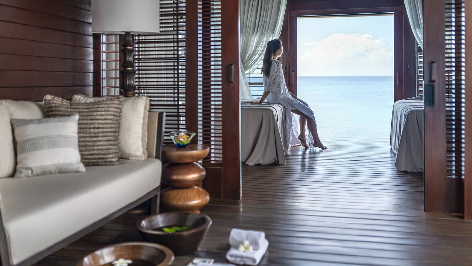 A woman leans on a massage bed on the far side of a spa treatment room and looks out through a large doorway onto the vast blue ocean