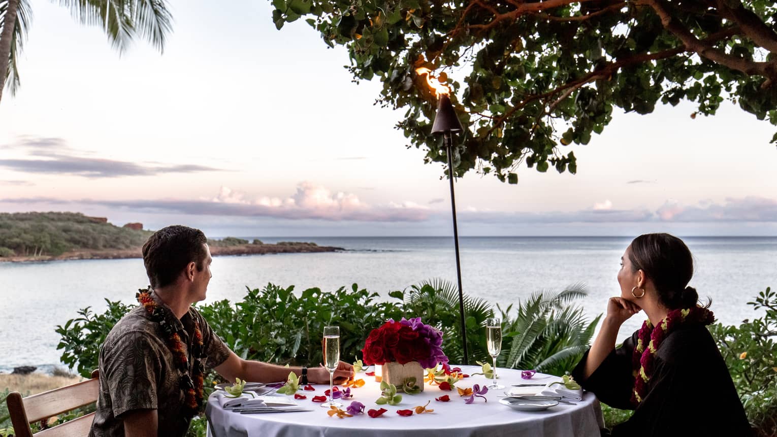 A couple wearing leis at a white table, and looking out on the ocean.