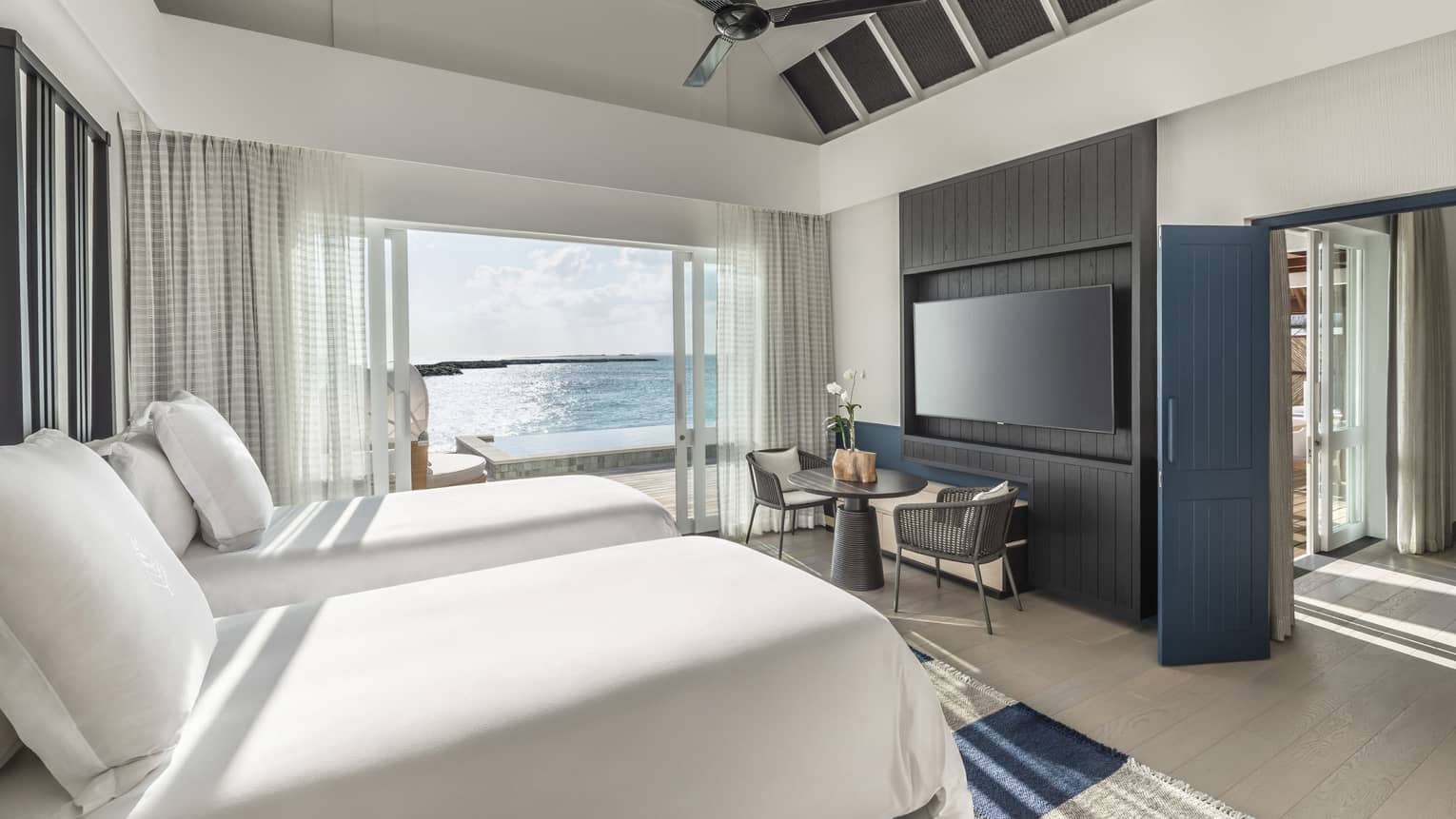 Bedroom with double beds, wall-mounted flat screen TV, two-seater dining table, doorway leading to pool deck