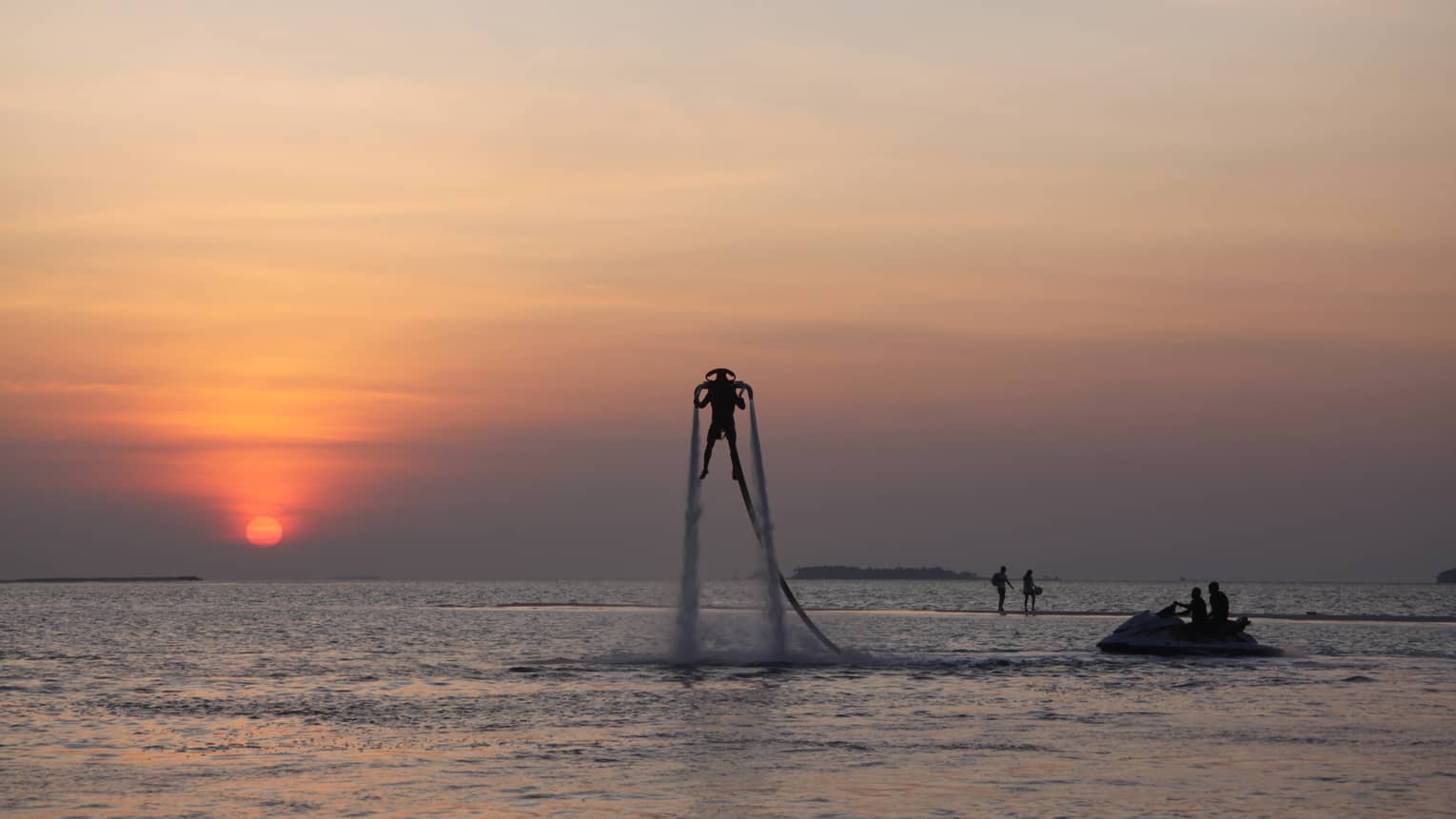 Silhouette of someone with X Jetpack suspended in air over water at sunset