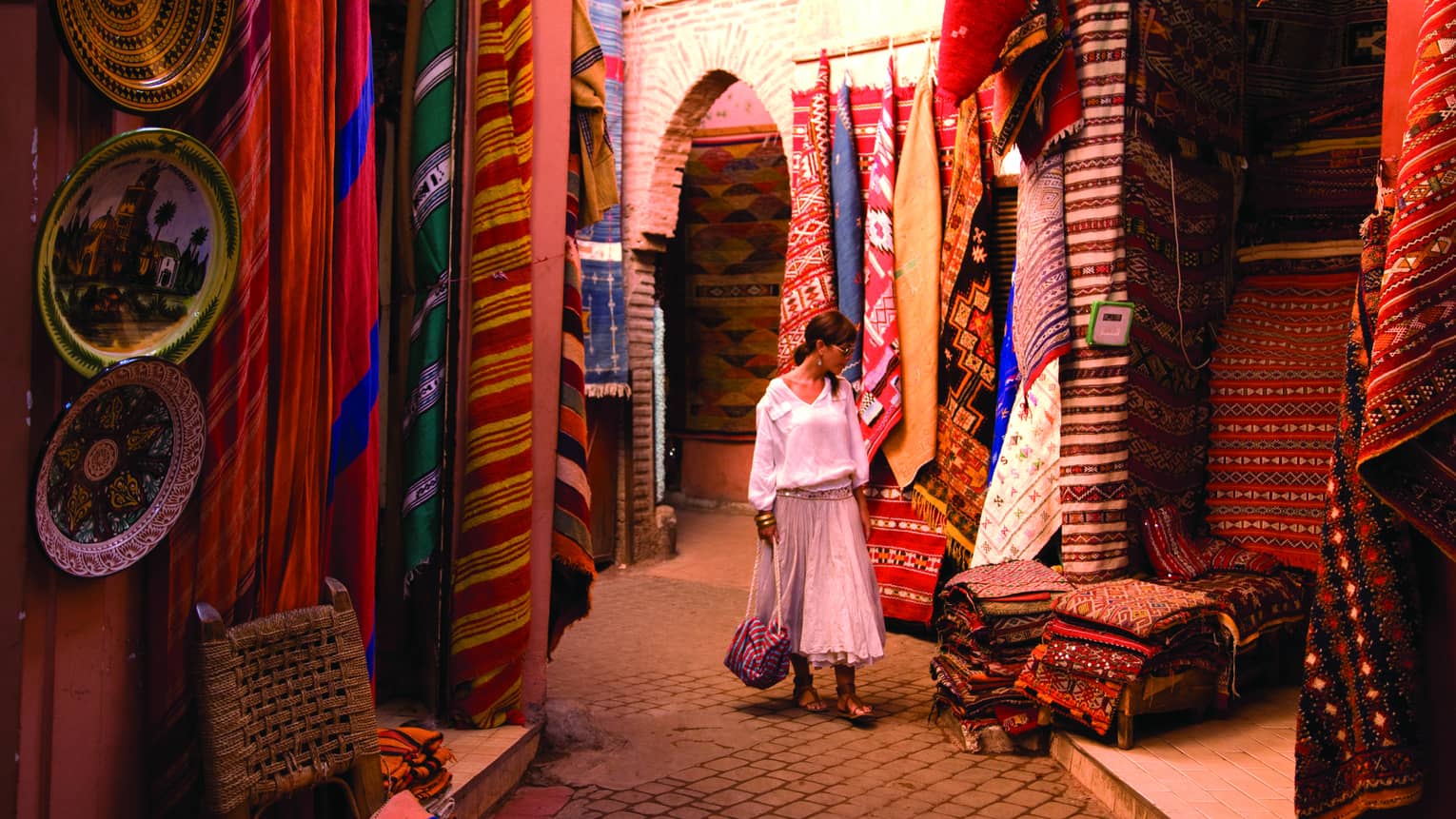 Woman holds purse, walks past hanging rugs, stacks of textiles in outdoor market