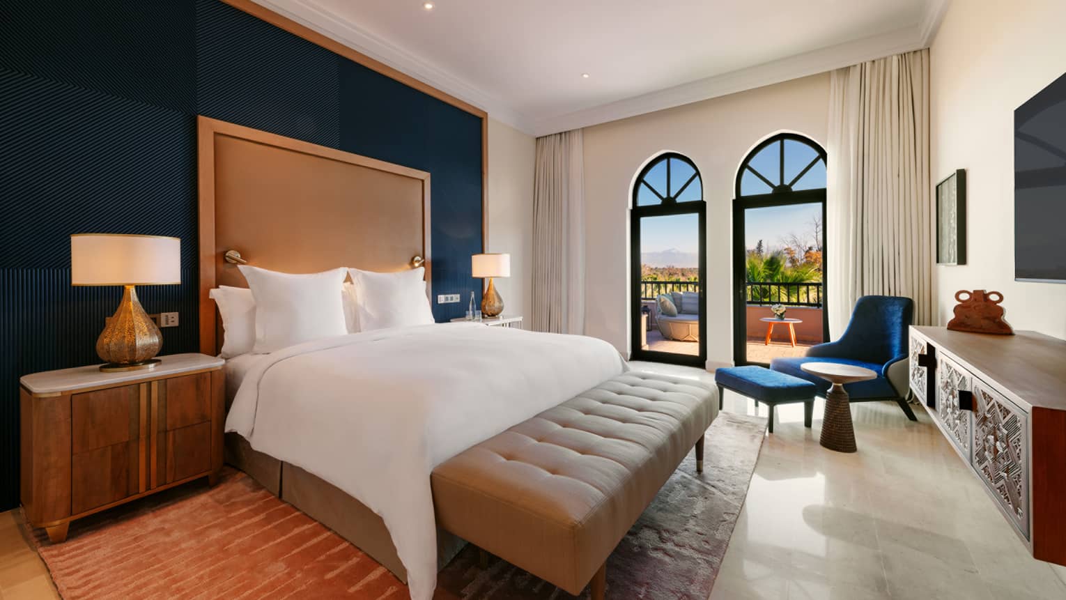 Luxury hotel bedroom with king bed, light brown headboard and bench, blue arm chair and ottoman in the corner, and walk-out terrace
