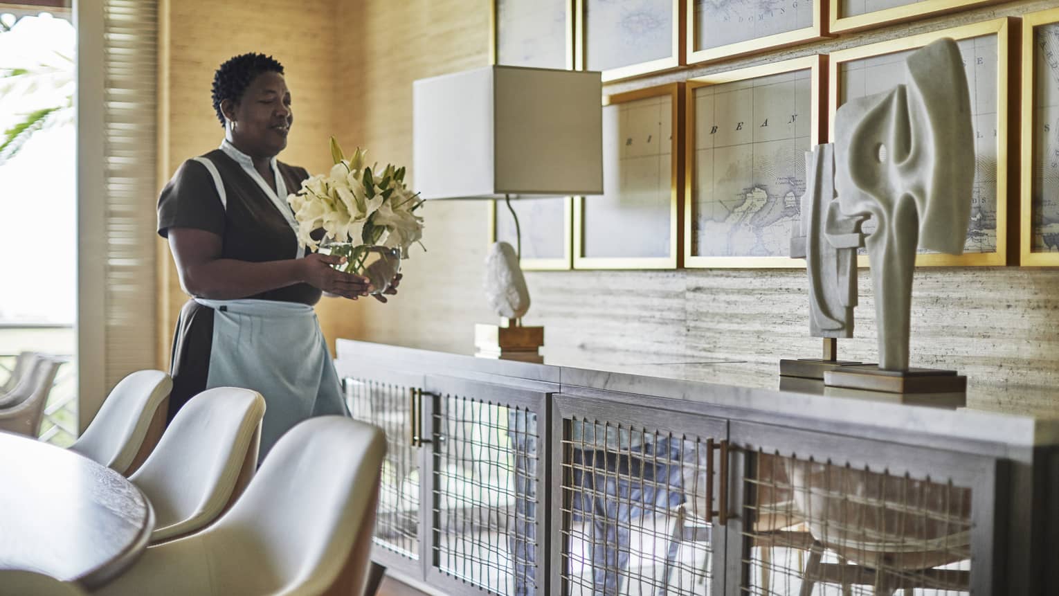 Housekeeping staff sets flower arrangement on dining room sideboard with lamp, modern sculpture