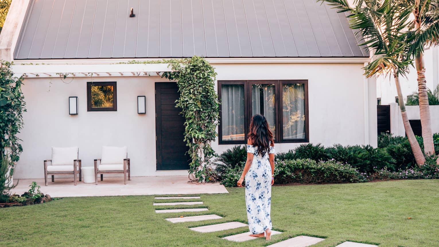 Woman walks along stone pathway in lawn to white house, palm trees