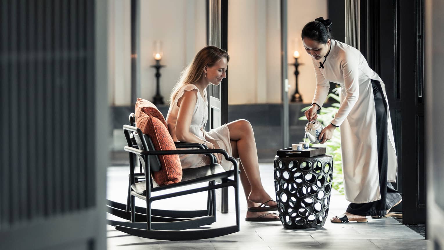 Server pours tea on tray on decorative black table for woman in rocking chair