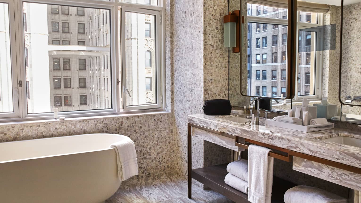 Luxury hotel suite bathroom with a freestanding bathtub, marble double vanity with mirrors and large windows overlooking city buildings