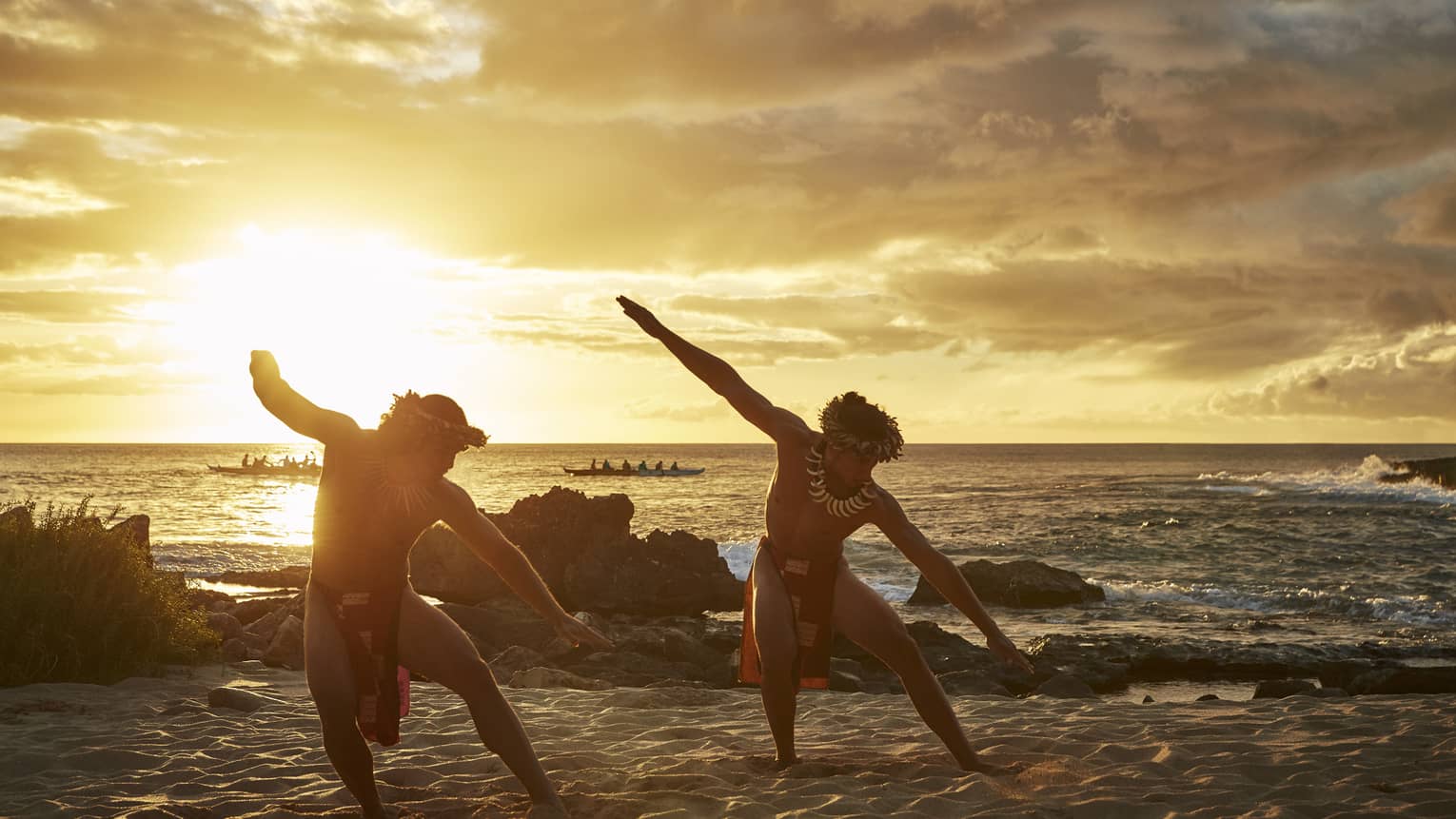 Two Hawaiian dancers with arms outstretched pose on sandy beach at sunset