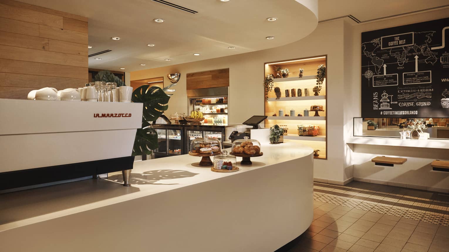Curved white counter with a white espresso machine on top and a small display of pastries, with another display and shelving on the other side of the room