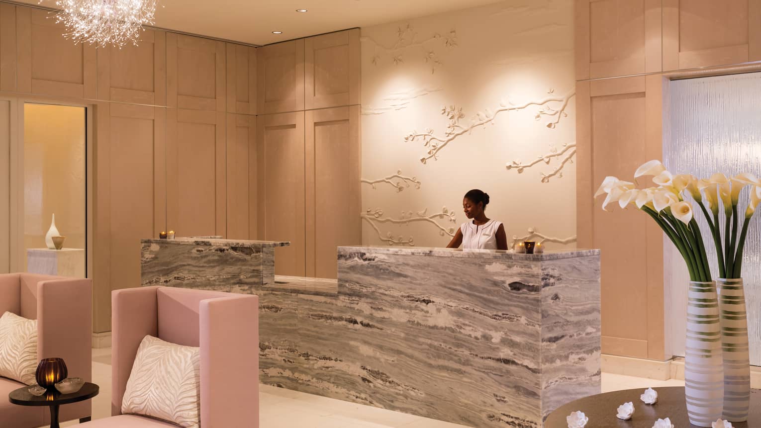 A well lit lounge of a hotel with light pink chairs for people to rest and a worker behind a grey and white desk area.
