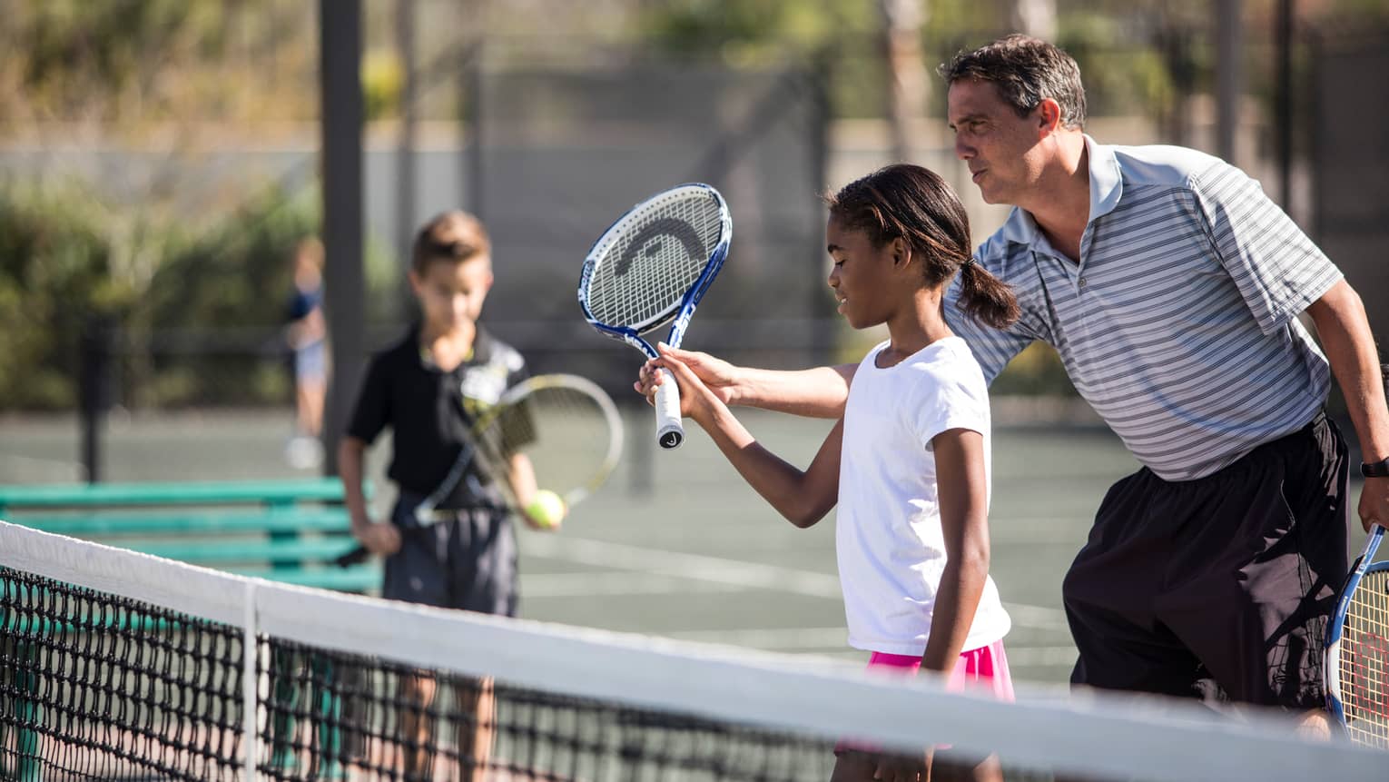 Tennis coach helps young girl hold tennis racket by net as young boy watches