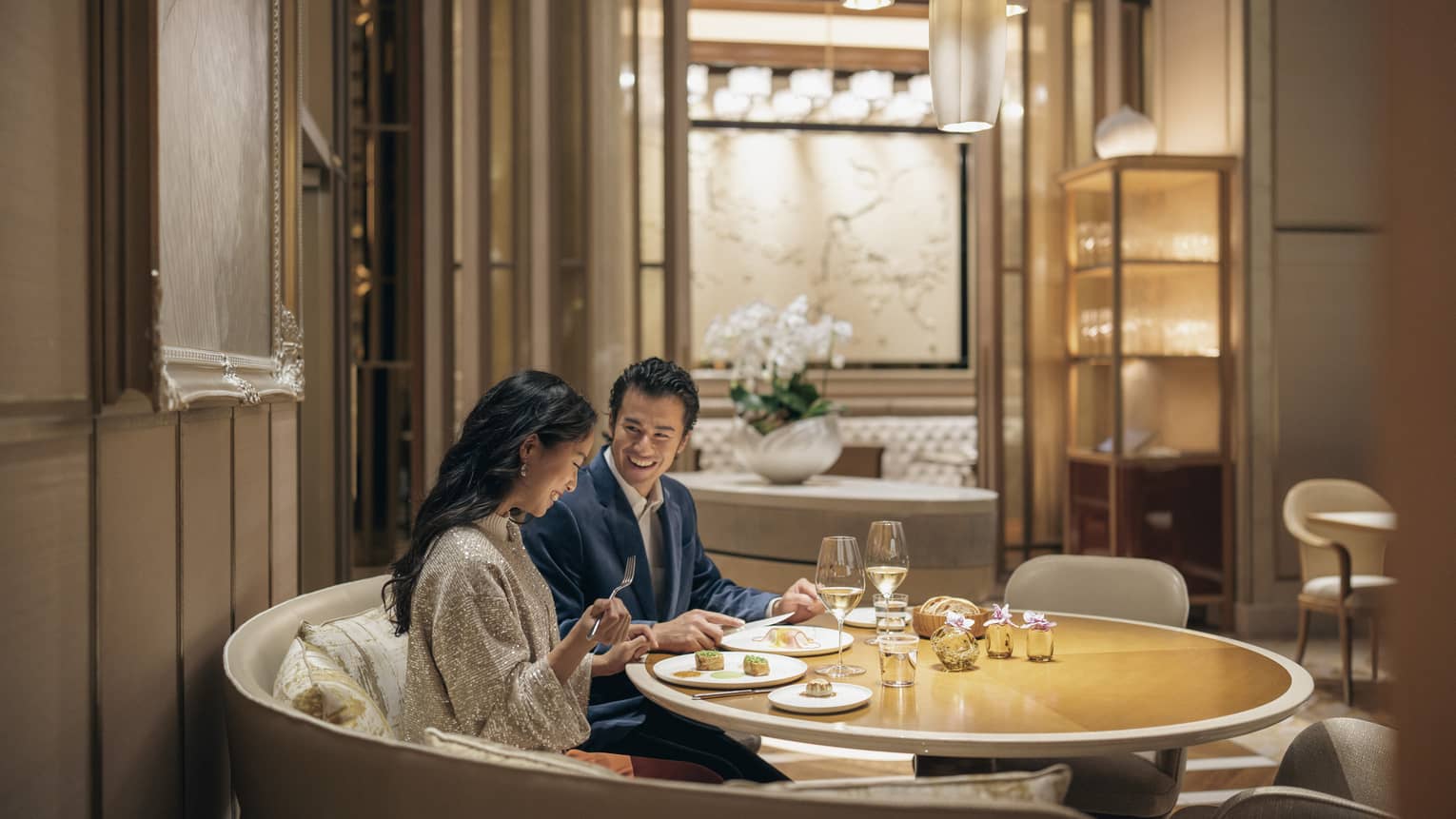 A smiling couple savours lunch with wine in a circular booth at a fine dining restaurant with warm pendant lighting.