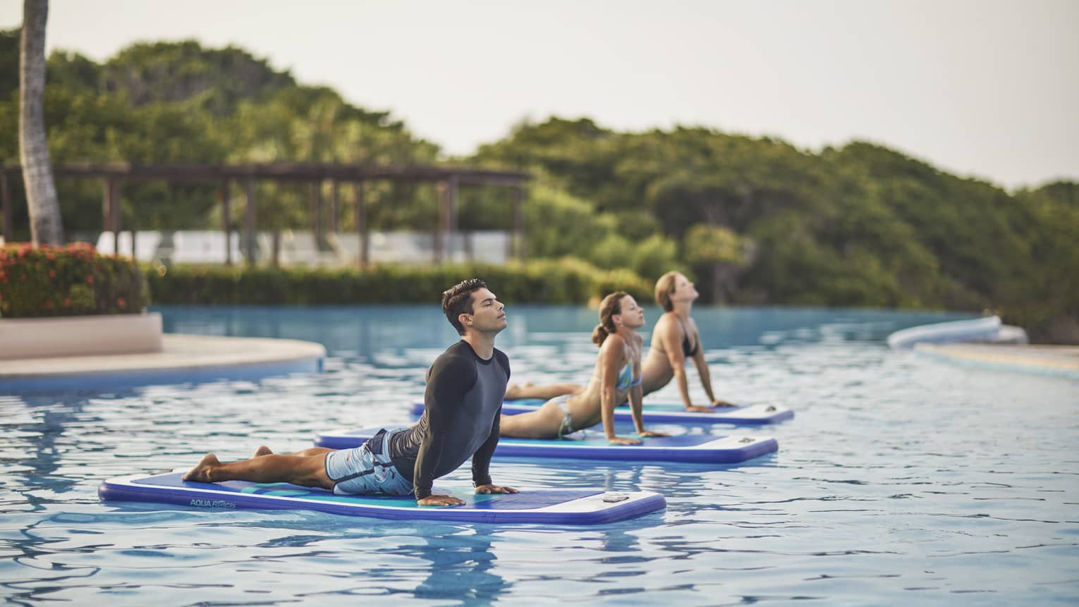 Man, two women in yoga poses on paddle boards on swimming pool