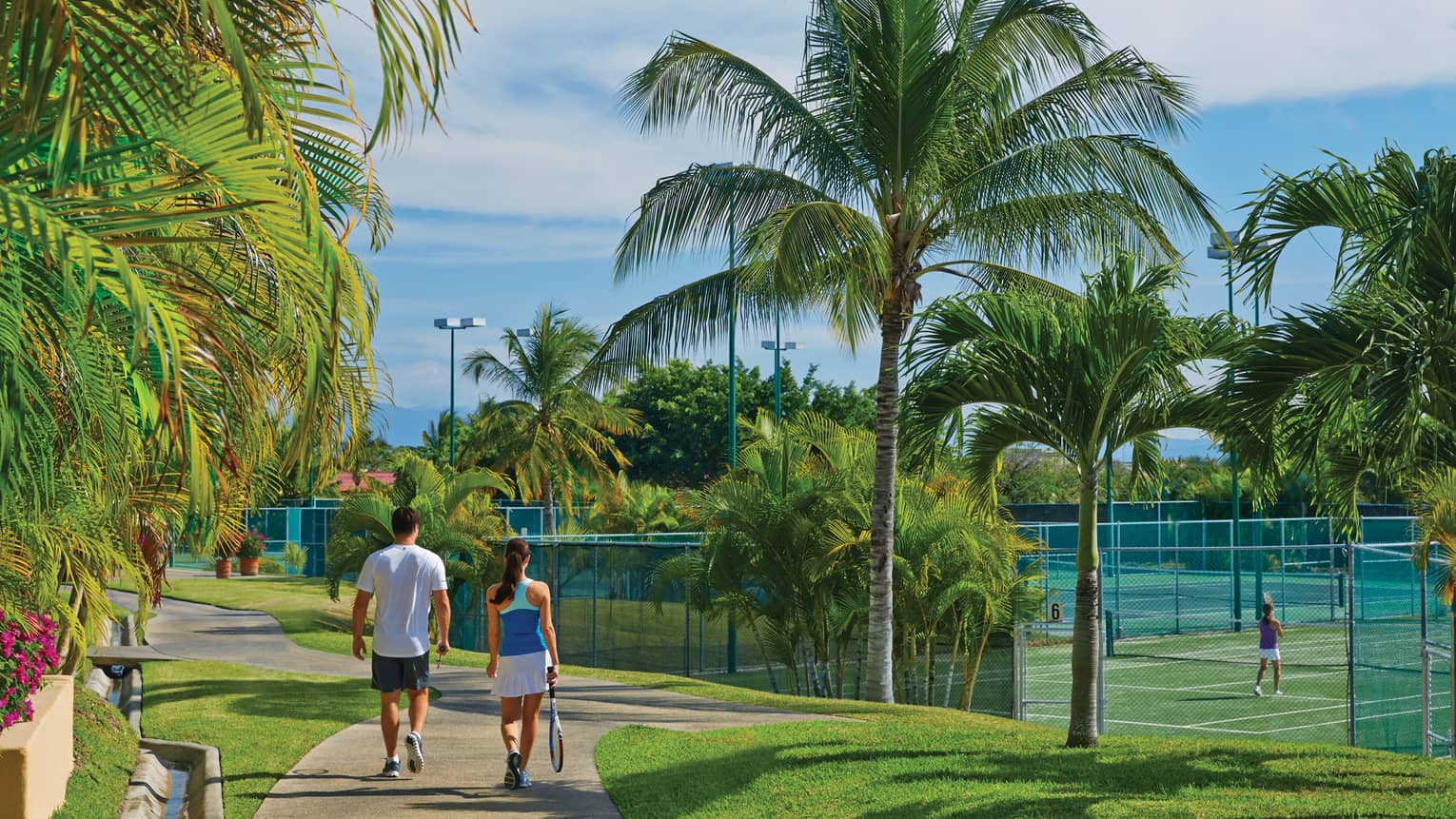 A man and woman walking along a path next to tennis courts and palm trees.
