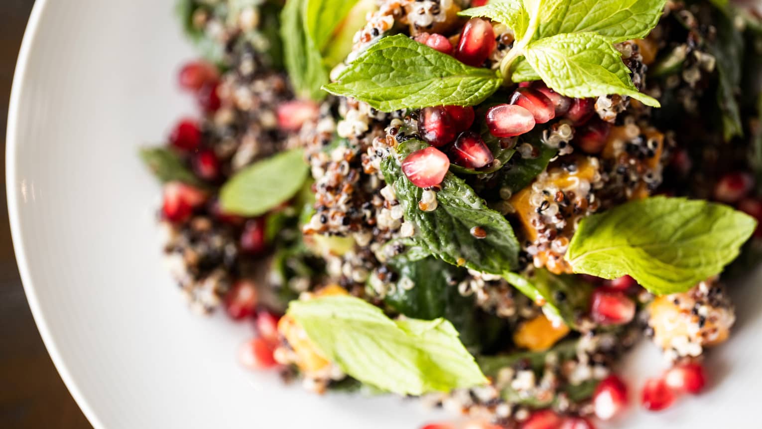 Fresh quinoa salad with green mint leaves, red pomegranate seeds and various vegetables all on a white round plate