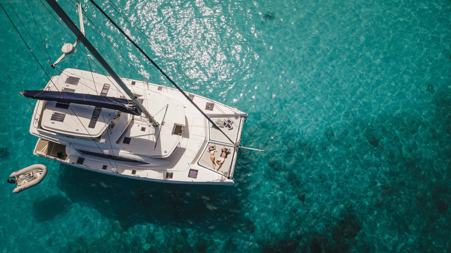 Aerial view of two sunbathers aboard a catamaran surrounded by clear, teal sun-dappled water rippling over rock formations.