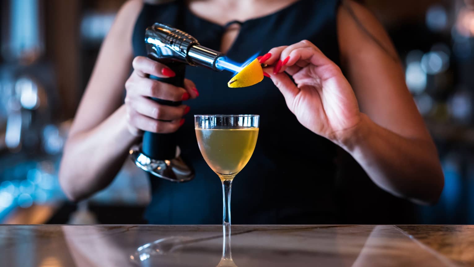 A woman using a blowtorch on an orange slide while a cocktail sits on the bar in front of her.