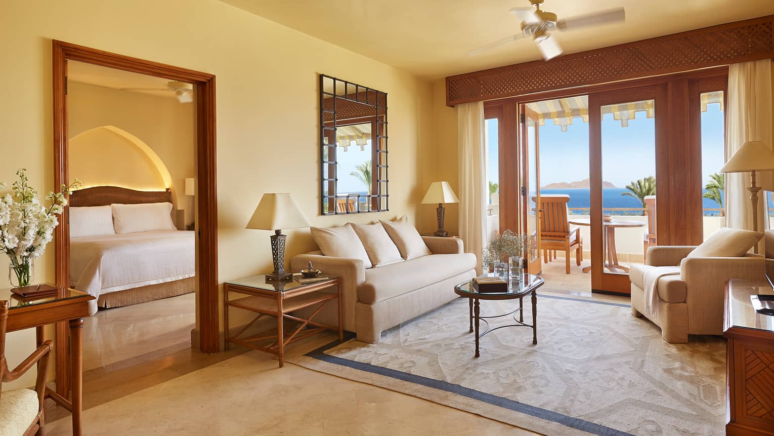 Hotel room seating area with white sofa, armchair, area rug, open balcony door, door to bedroom