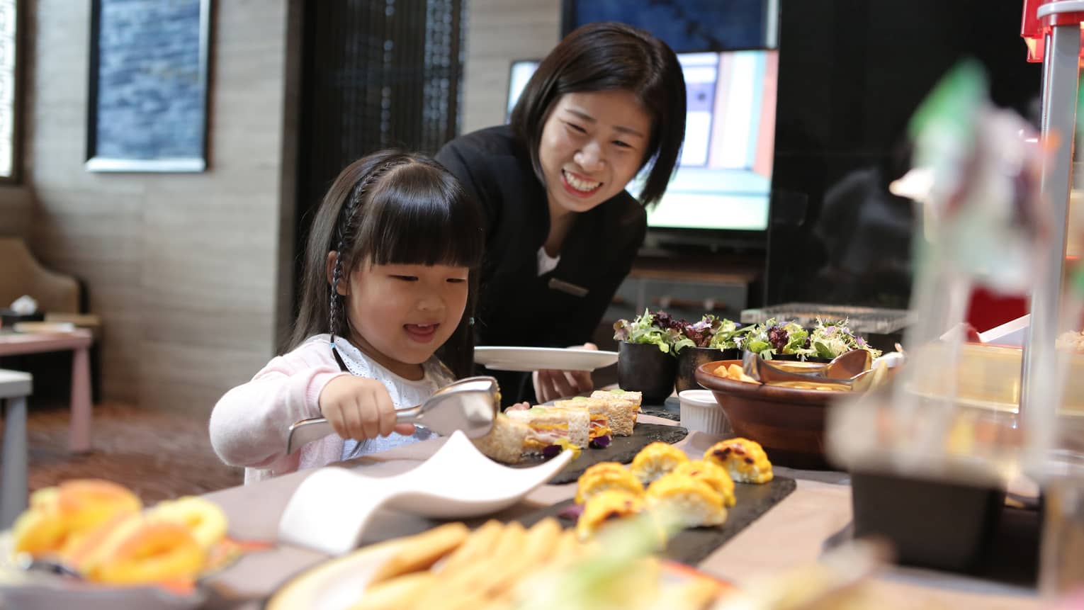 A little girl chooses items from the kids buffet while a smiling staff member looks on