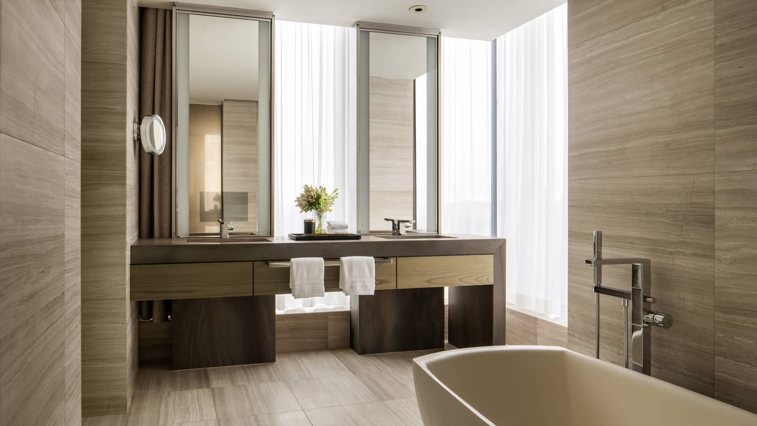 Luxury hotel suite bathroom with light wood paneling, a freestanding tub and a dual-sink vanity under large windows 