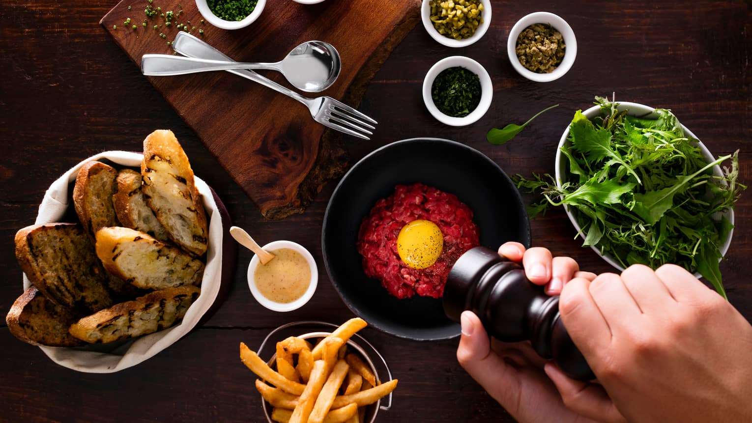 Hands grinding pepper over bowl of tartar, baskets with garlic toast, fries, lettuce, spices
