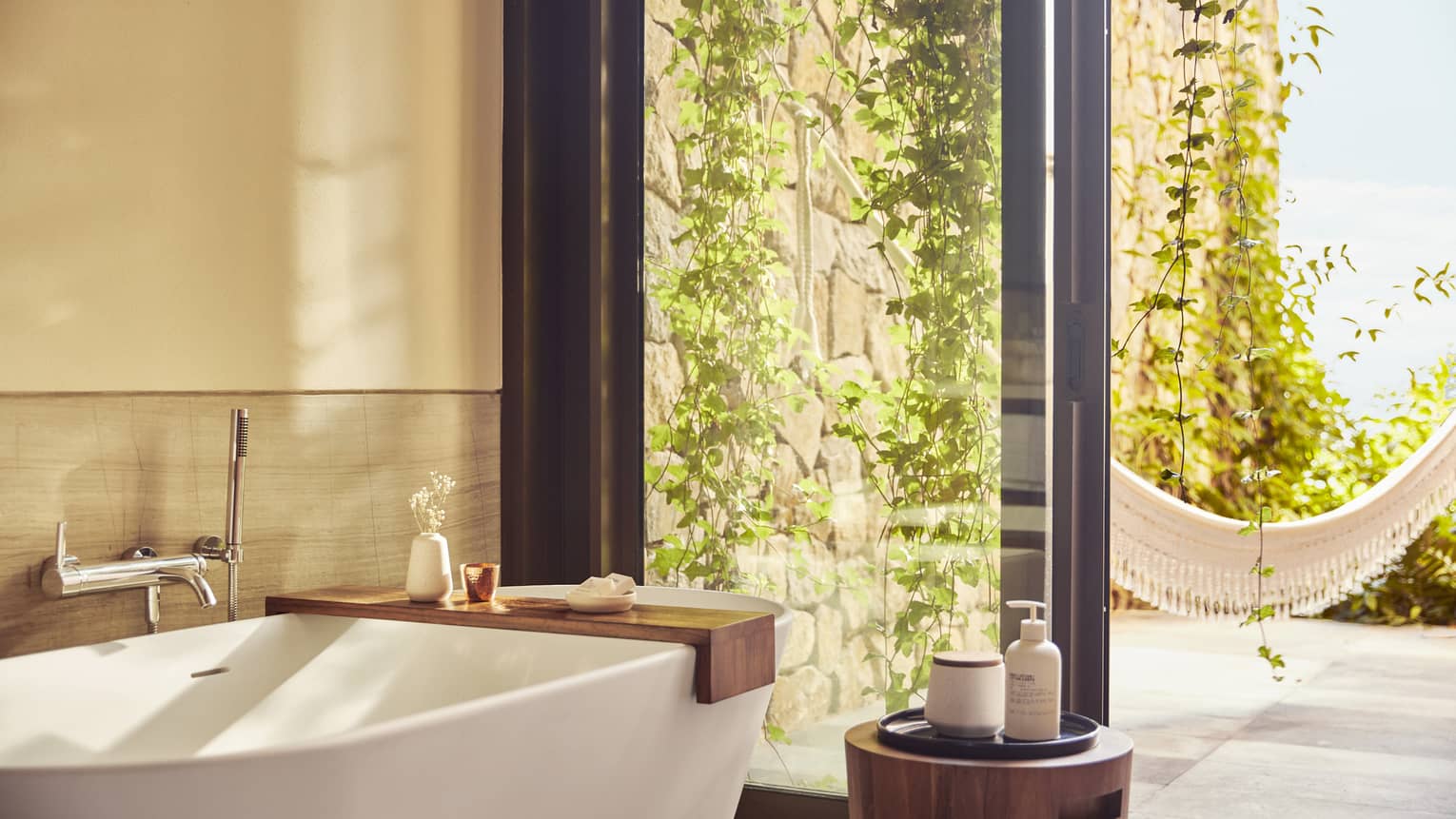 Luxurious bathroom with a free-standing tub, large glass doors and a view of an outdoor hammock. Lush vines climb the stone wall, bringing nature into the serene space.