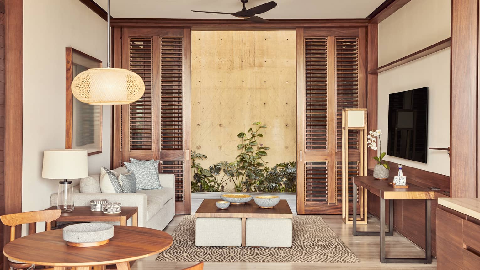 Cozy living area with a grey sofa, wooden furniture and a small dining table. The room features large shutters, soft lighting and indoor plants, creating a warm, inviting atmosphere.