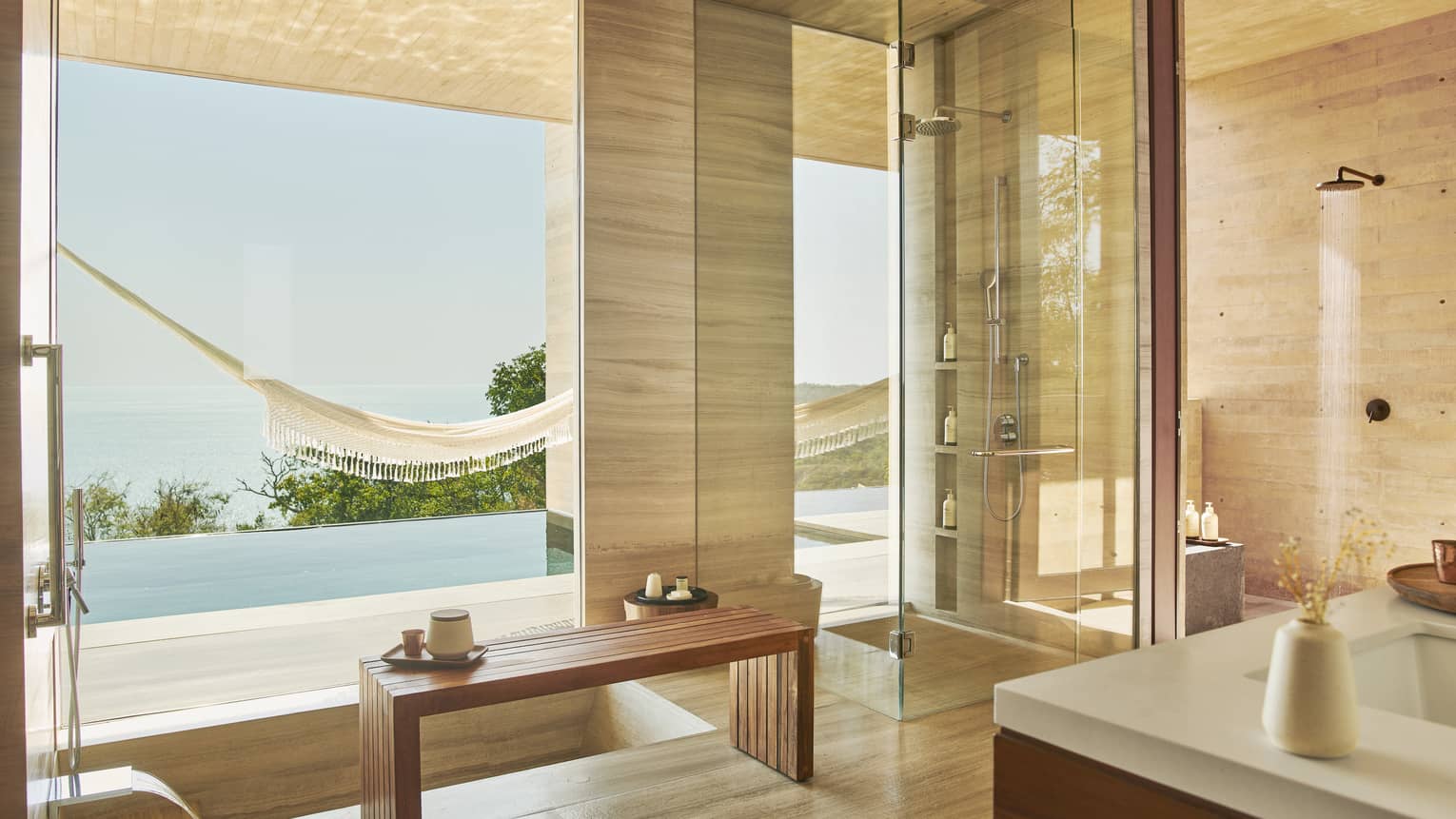 Luxurious bathroom with a glass-enclosed shower, wooden accents and a large window overlooking a hammock, infinity pool and serene ocean view, creating a tranquil atmosphere