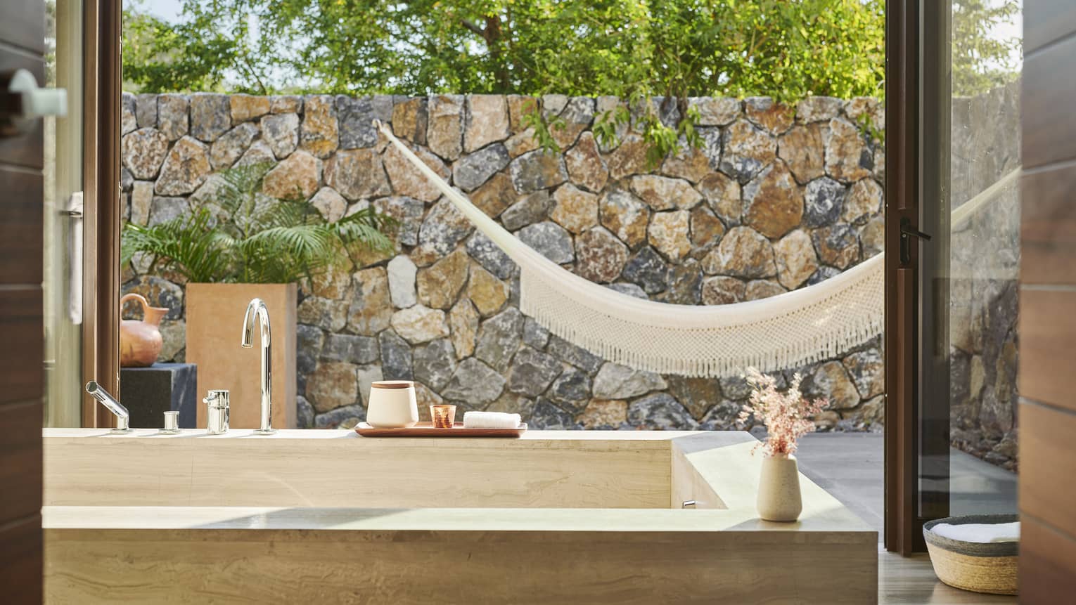 A luxurious bathroom with a stone bathtub facing an outdoor area featuring a hammock, stone wall and greenery. Natural light pours through open glass doors, creating a tranquil setting.