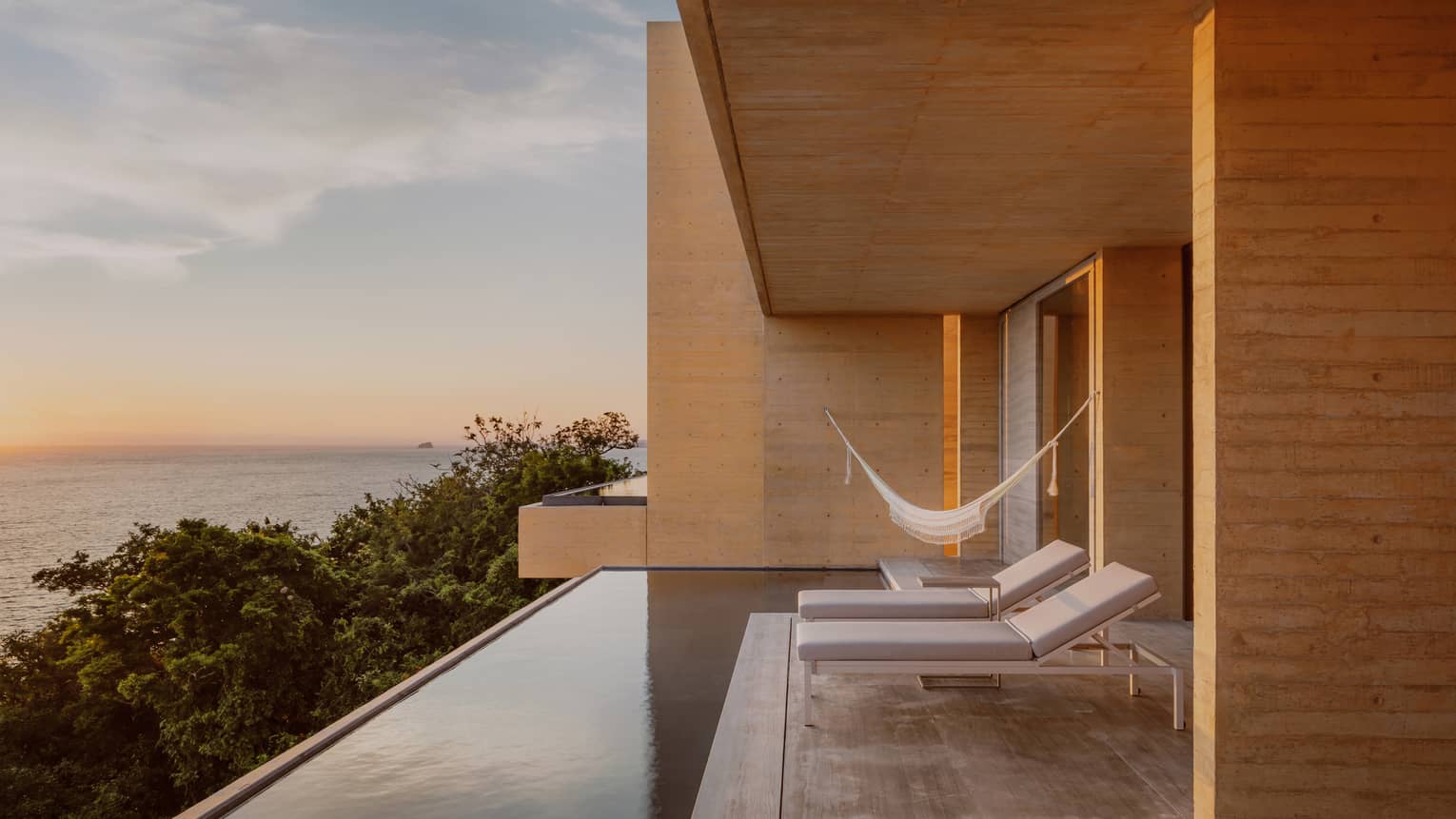 Modern terrace with lounge chairs, a hammock and an infinity pool, overlooking lush greenery and the ocean at sunset. The space features minimalist wooden architecture and a serene atmosphere.