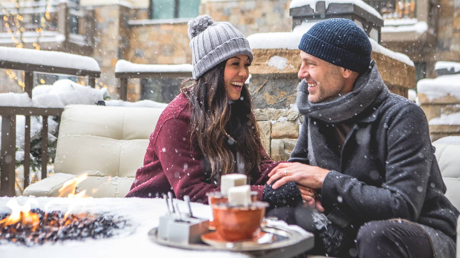 Smiling couple sitting by a firepit in a snowy outdoor setting, bundled in winter coats and hats. Snowflakes fall around them as they enjoy a cosy moment. 
