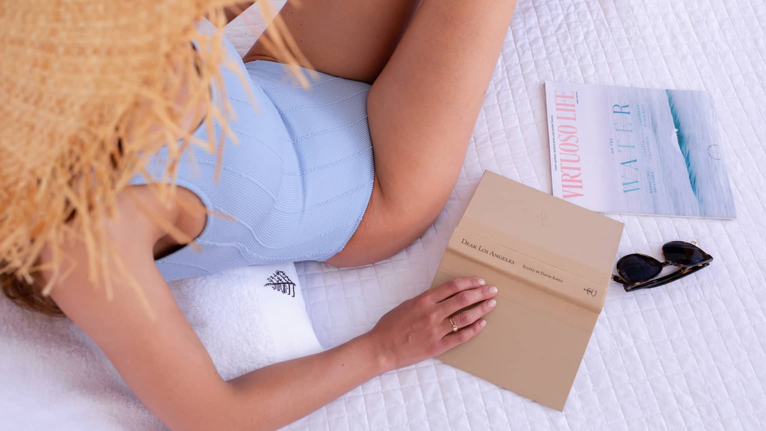 A woman in a blue swimsuit and straw hat holding a tan book while lounging on white bed