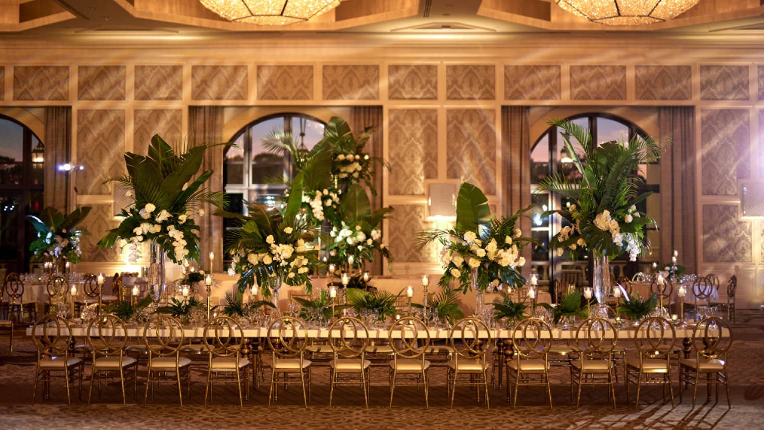 Ballroom with row of arched windows set up for a wedding reception with a long rectangular table topped with flowers, greenery and candles