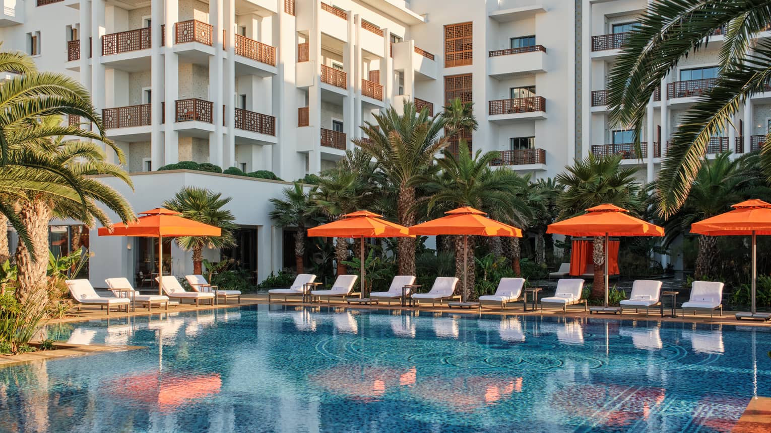 Pool lined with orange umbrellas by white lounge chairs outside of white building and surrounded with palm trees