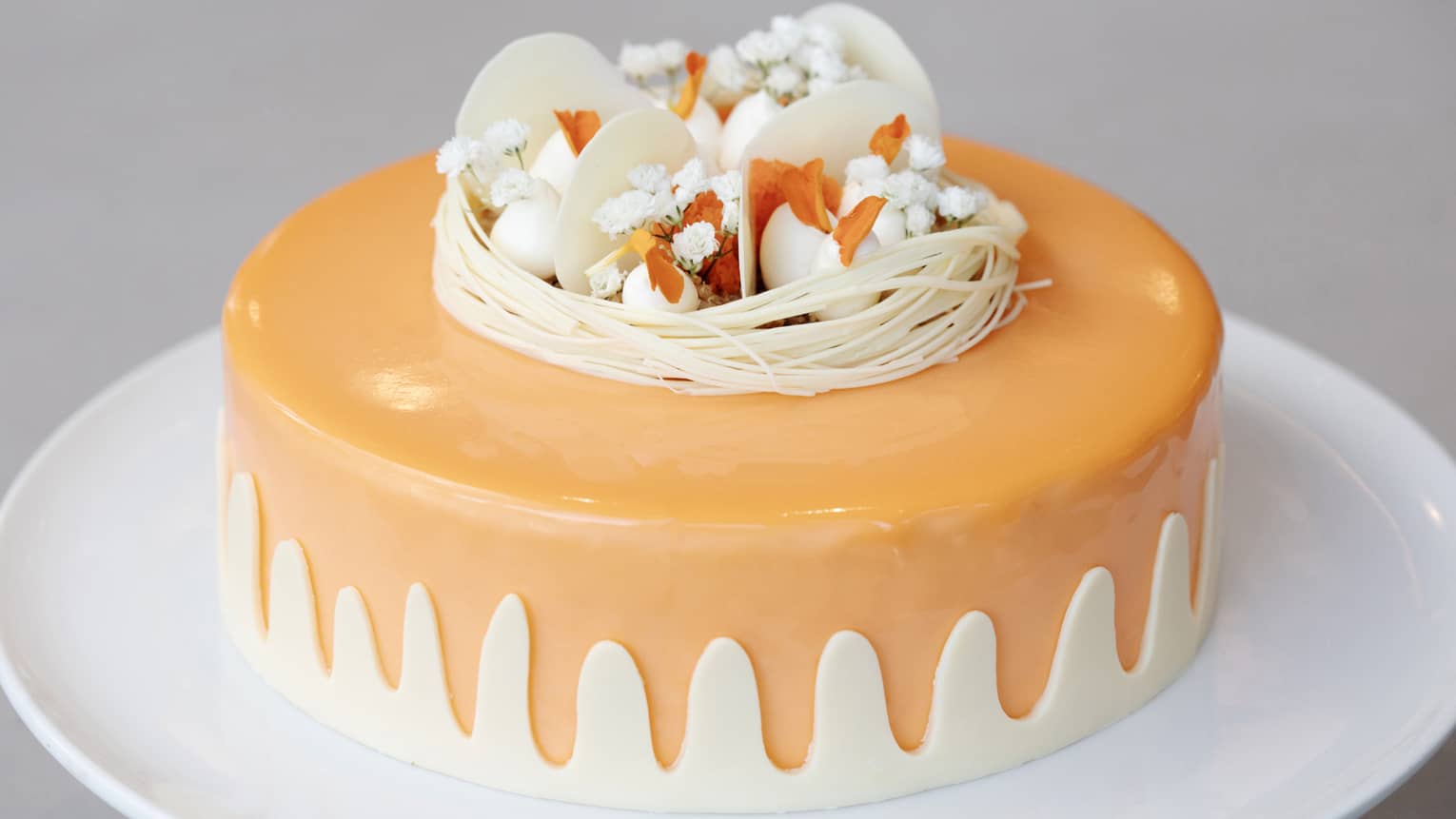 A white plate holding a smooth orange cake that has a white scalloped decorative edge around the bottom and is topped with white garnishes and decorations in various shapes and textures