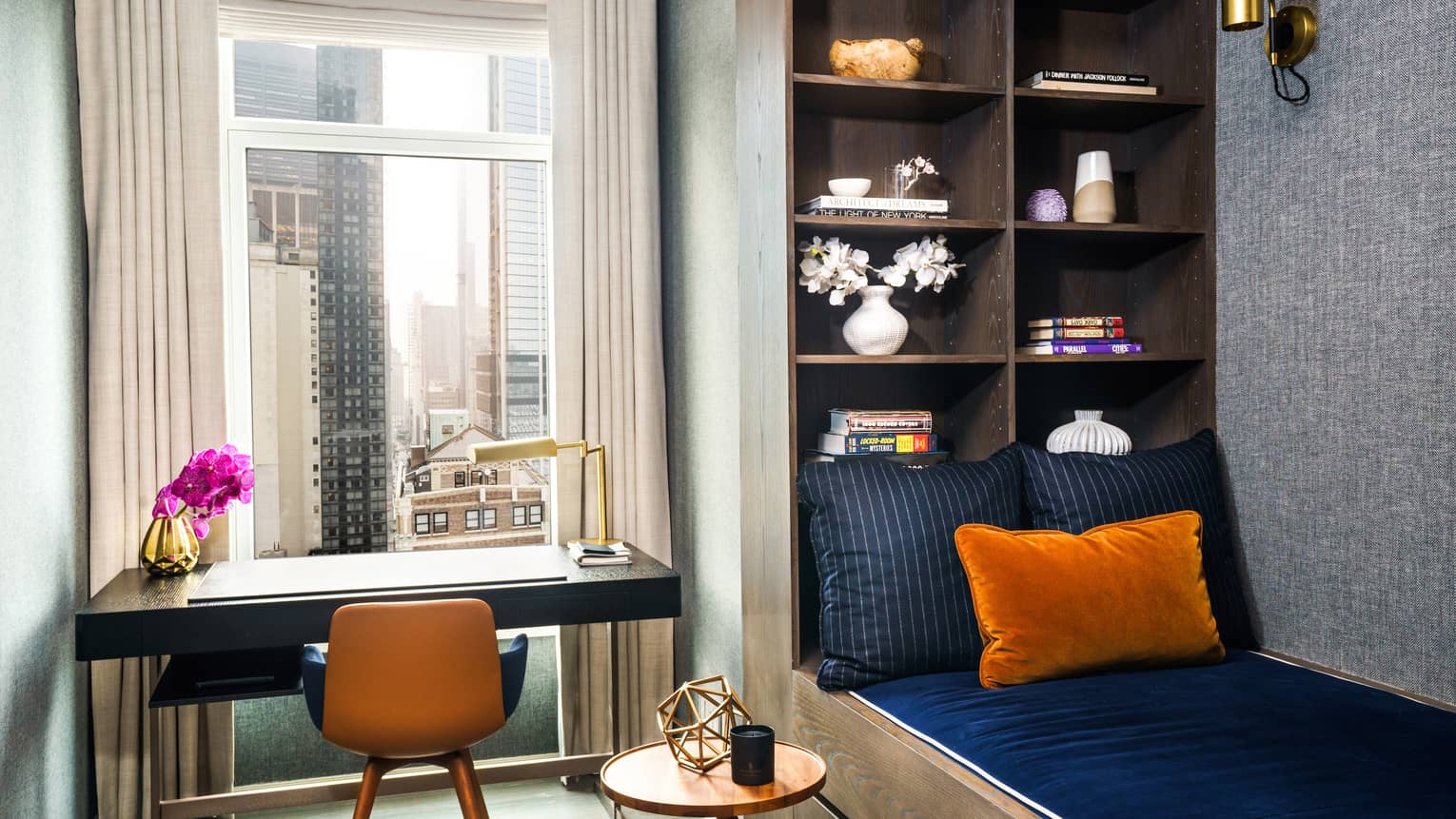 Twin bed with dark blue linens, adjacent to wall with built-in shelves, writing desk, city view