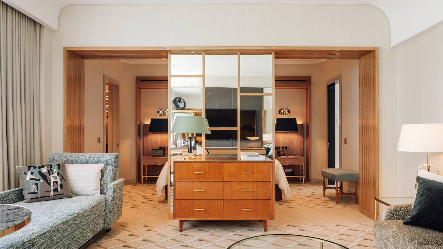 Hotel bedroom connects to living room via glass wall divider, with wooden dresser and chaise