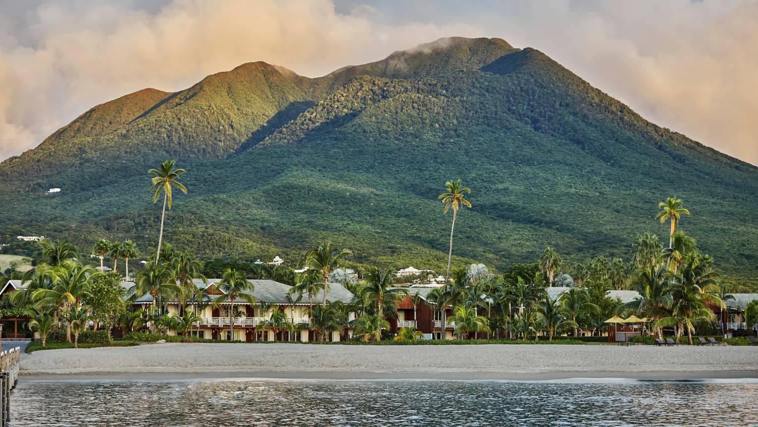 Buildings and palm trees on an ocean shore with a large mountain in the distance.
