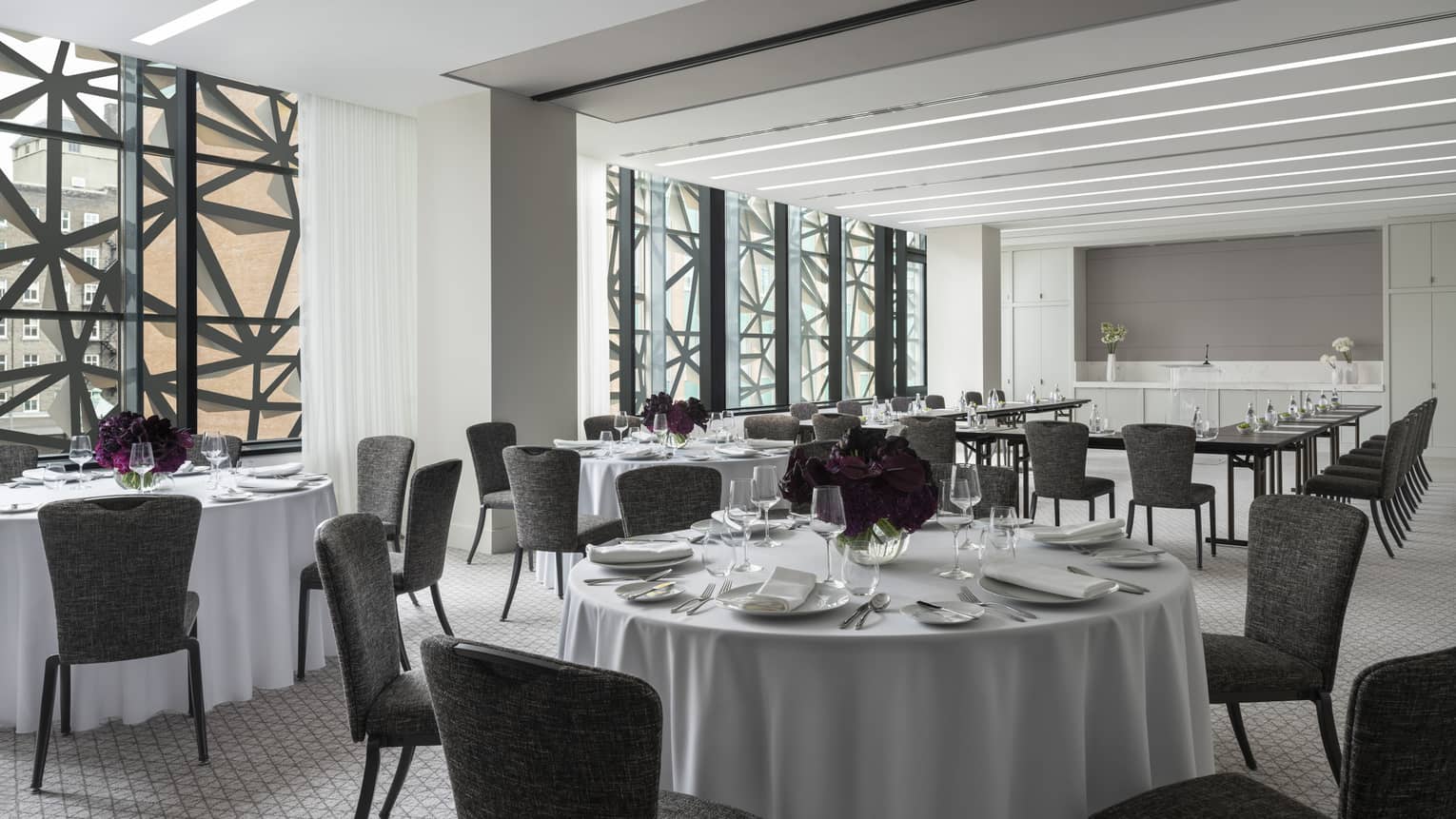 Salon with white round tables and flowers, grey chairs, U shaped table in background, ceiling-to-wall windows