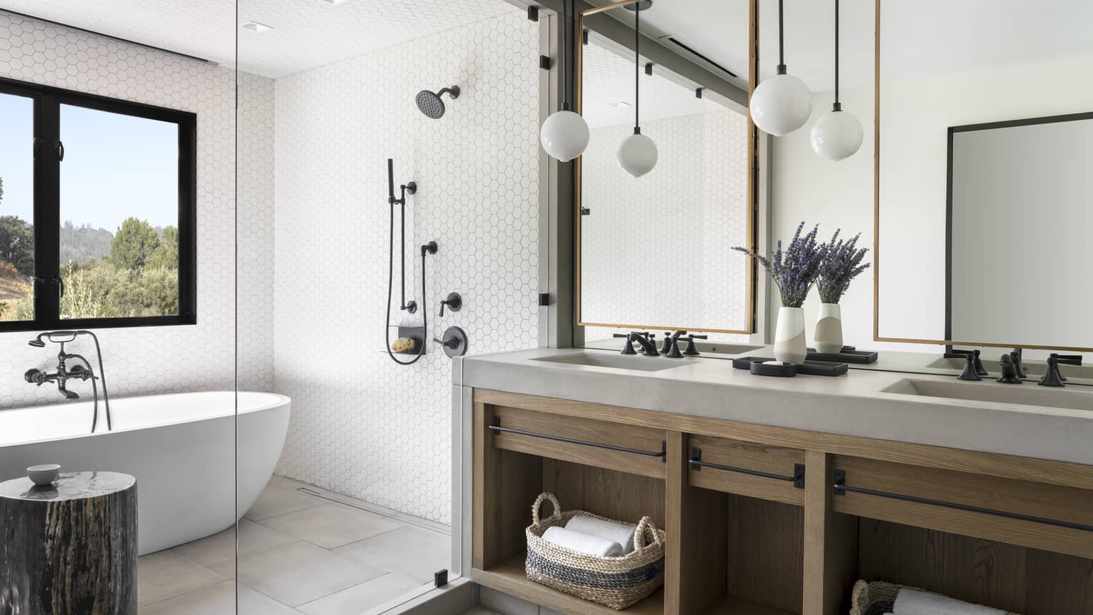 Luxury resort bathroom with a freestanding bathtub, walk-in shower, double vanity with wooden cabinetry, mirrors, hanging globe lights and lavender decor
