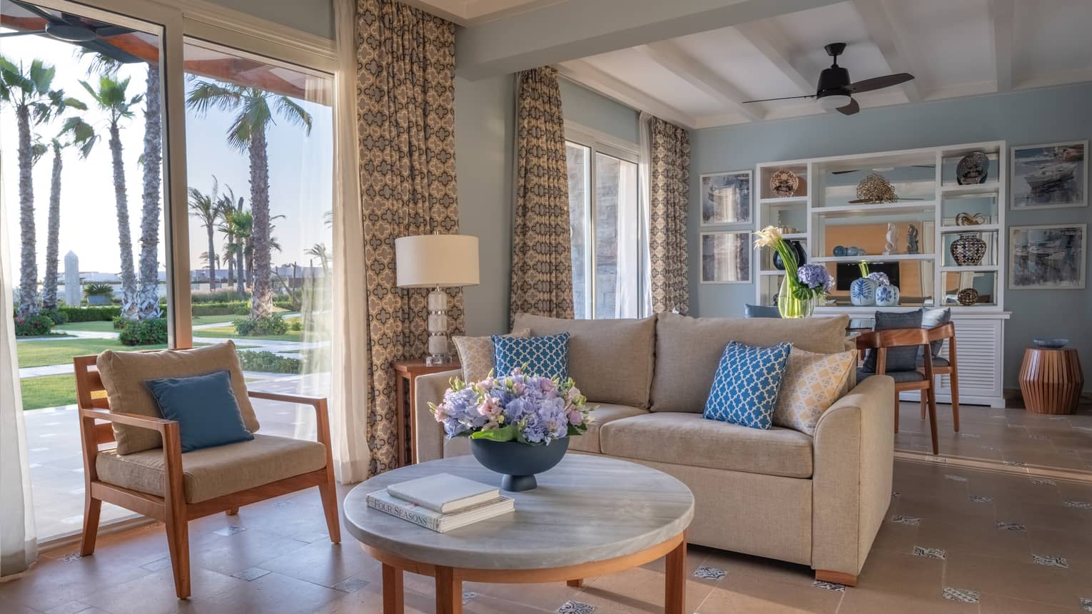 Guest room living area with tan sofa, armchair, round marble coffee table, dining table in background, sliding glass door to patio