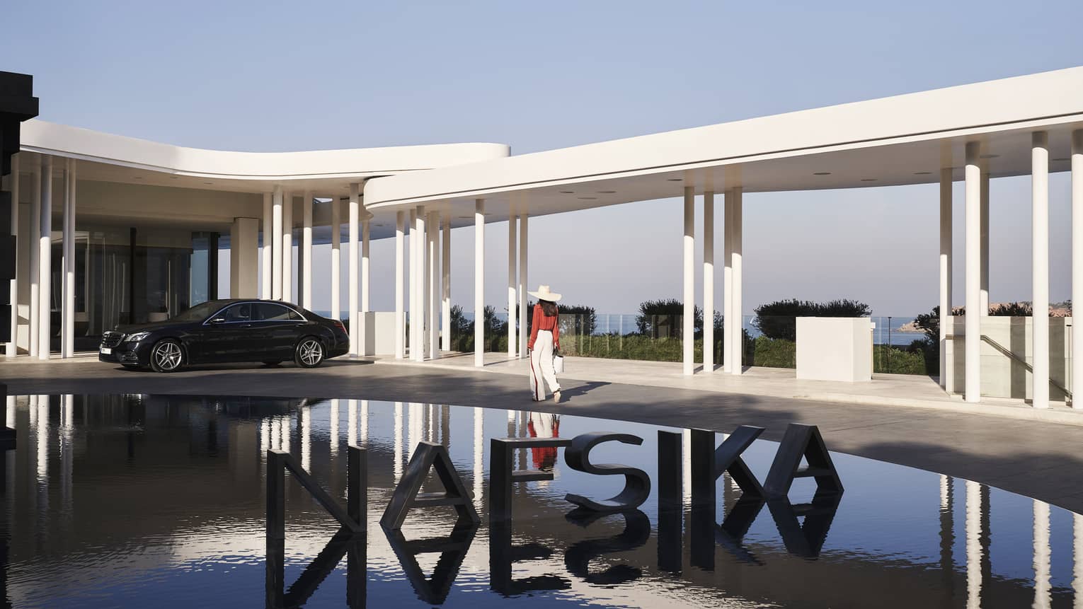 A guest passes a metal sign reading NAFSIKA in a fountain and towards an elegant car parked under modern white porticos.