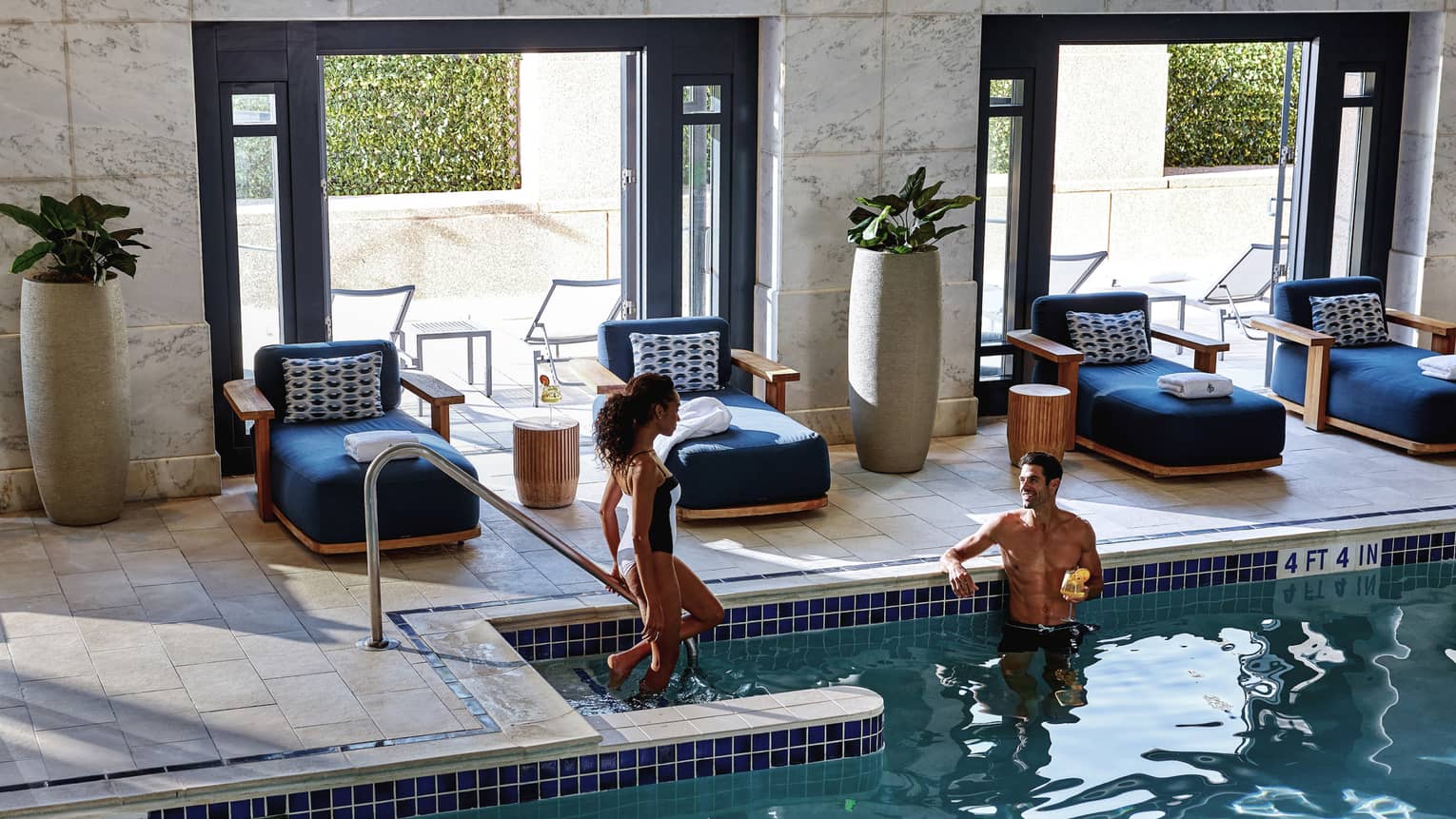 A woman steps down into an indoor pool towards a man lounging while holding a yellow cocktail