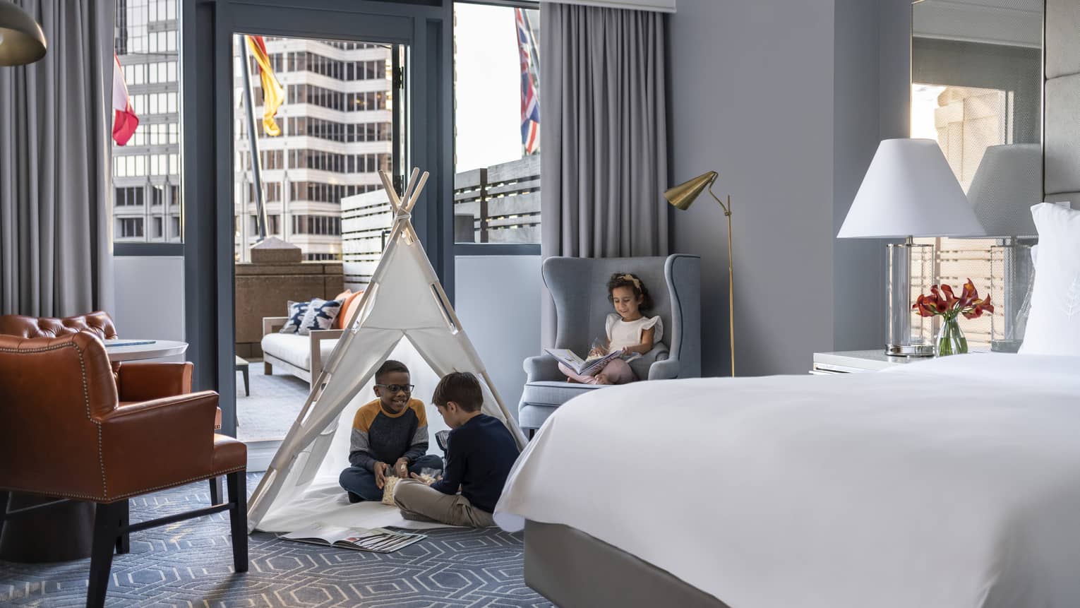 Children sitting in a tent and on a chair in a hotel room.