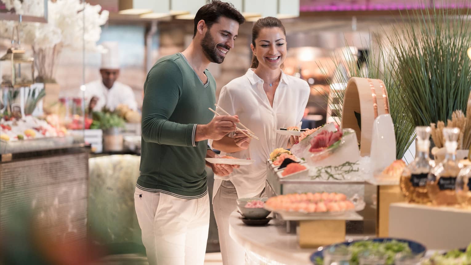 A man wearing white pants and green sweater stands next to a woman wearing all white at the sushi table of a buffet