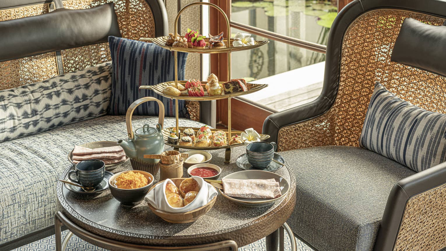A round table bordered by rattan furniture holds a stoneware tea set and a gold three-tier tray with sweet and savoury bites.