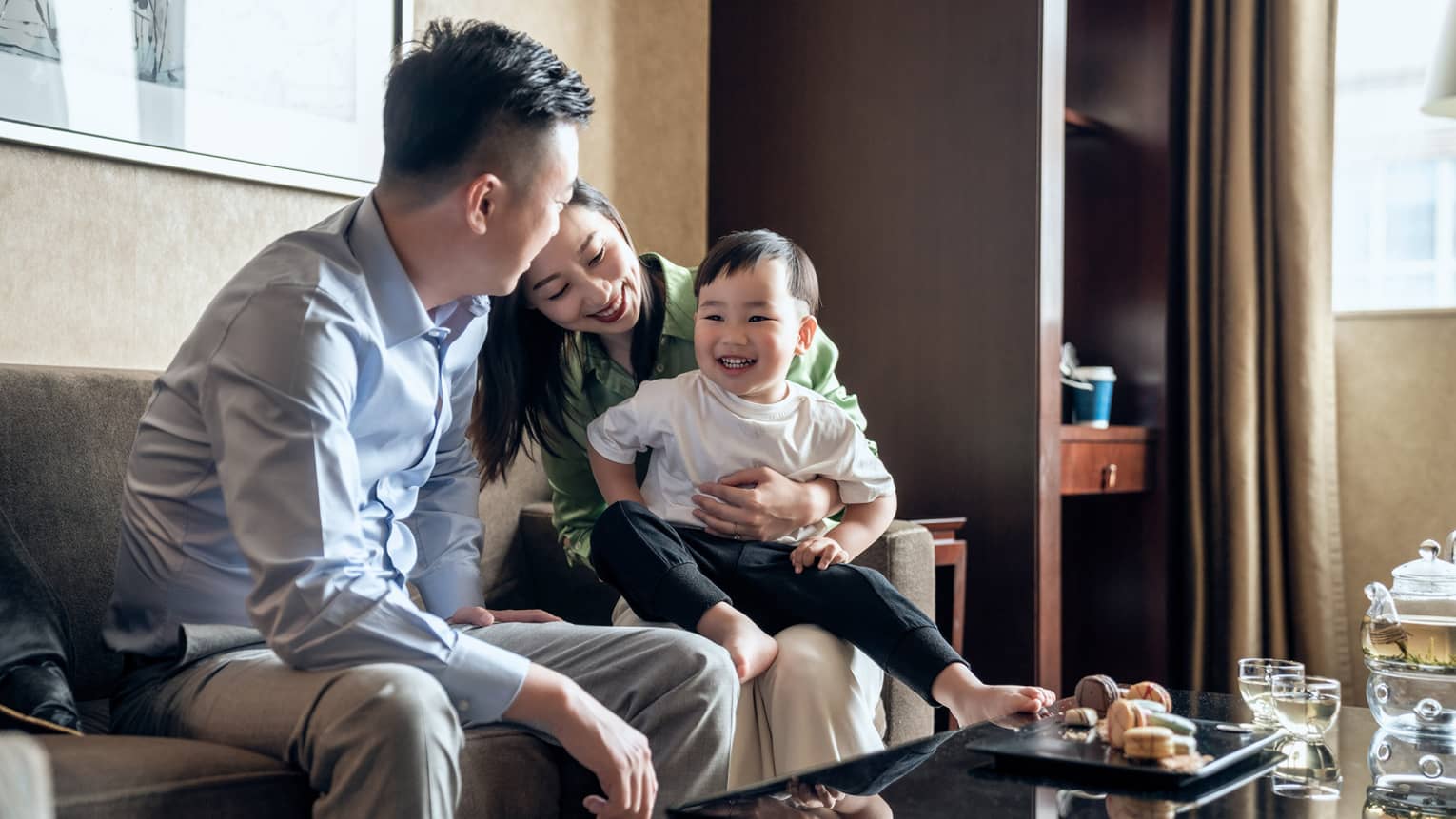 Laughing couple sits with their toddler son, tea and sweets on the coffee table in front of them
